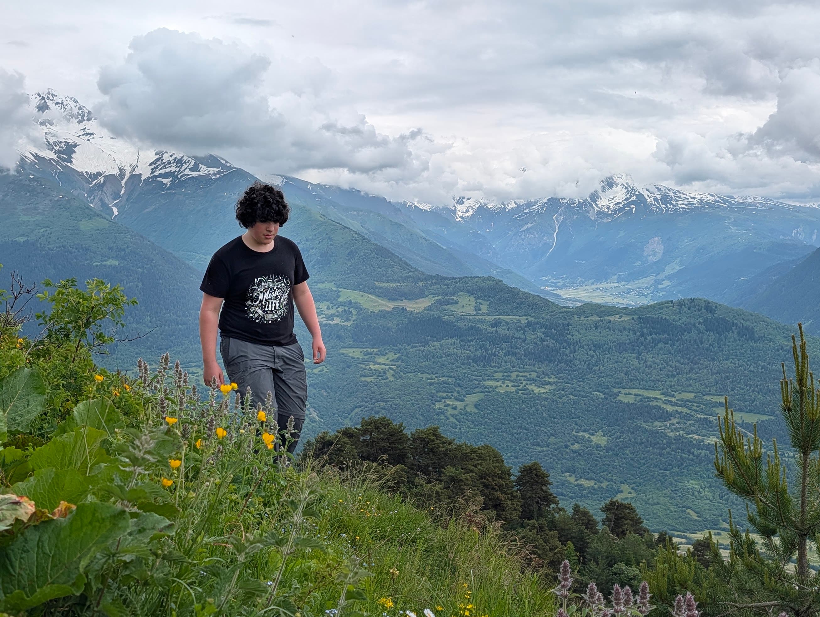 Orr Knaan walking a mountain path, with a wooded valley and snow-covered peaks behind.
